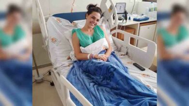 Deitada em um leito de hospital, usando jaleco verde, Giovanna se mostra sorridente.