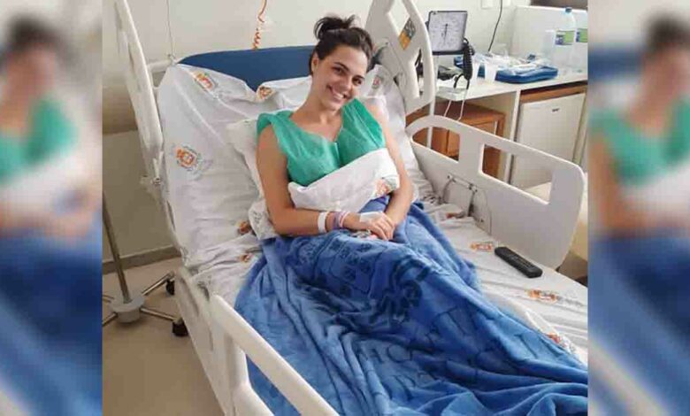 Deitada em um leito de hospital, usando jaleco verde, Giovanna se mostra sorridente.